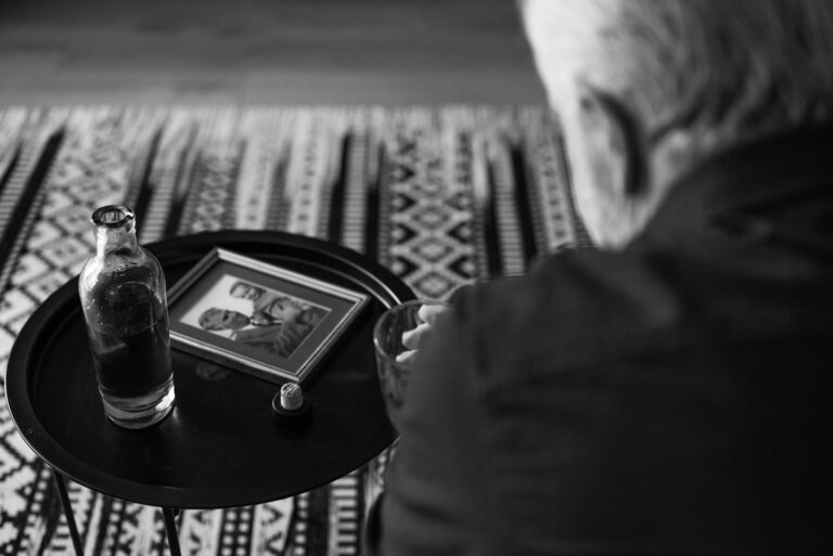 Black and white image of an elderly man reflecting with a framed photo and drink.