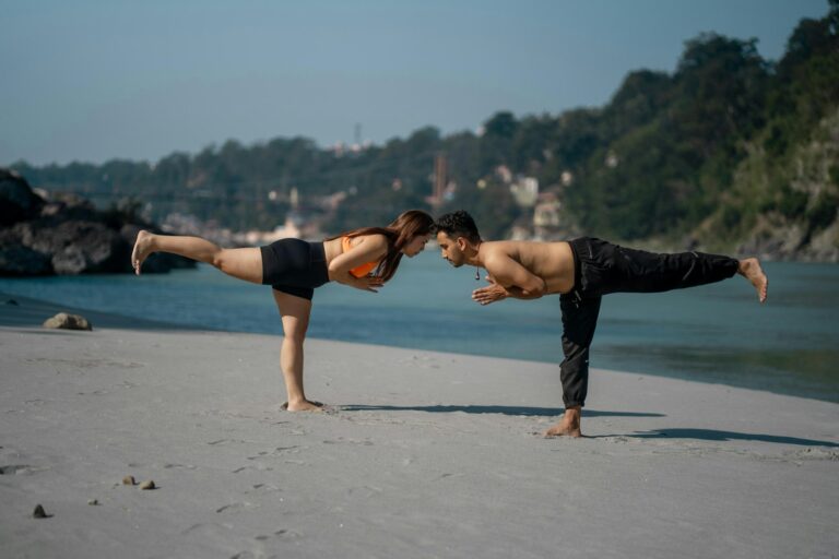 Couple doing yoga on a sandy beach by the water.